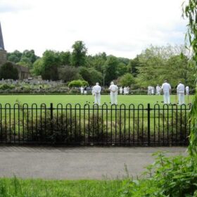 Wrought iron fence in foreground with bowling green behind and bowlers in 'whites' playing a game of bowls with trees in the background